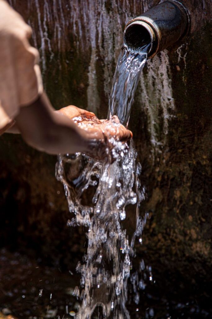 pexels photo 11612595 Close-up of hands collecting flowing water from a pipe outdoors.